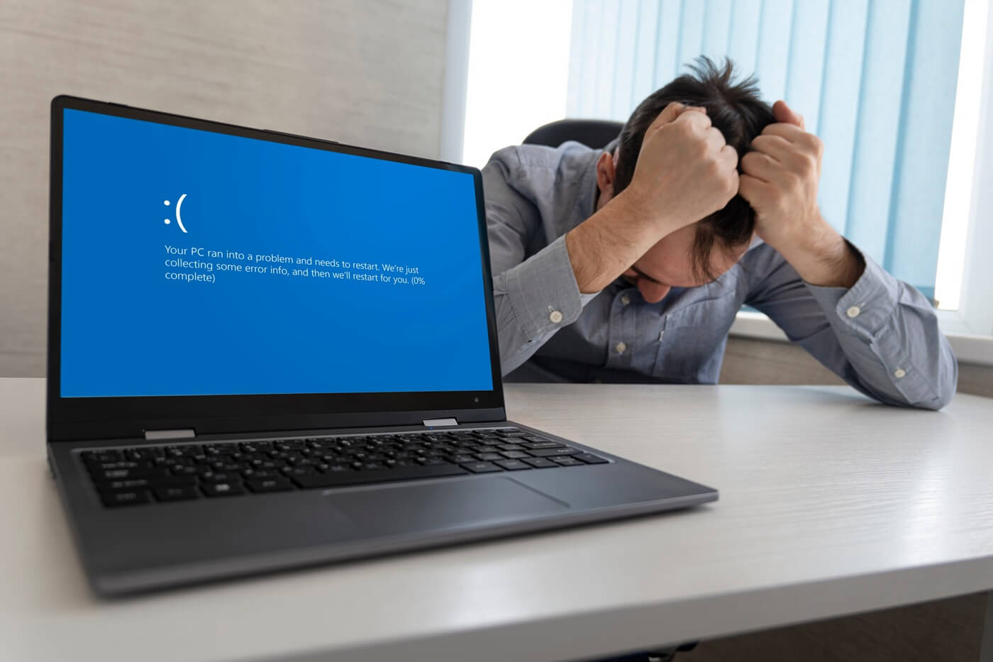 A desperate man clasps both hands over his head at the desk, and an error message from Windows is visible on the laptop screen.