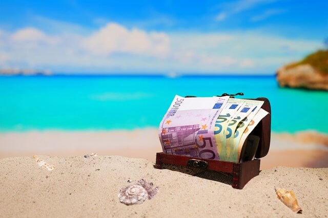 A small treasure chest filled with banknotes on the beach with the sea in the background