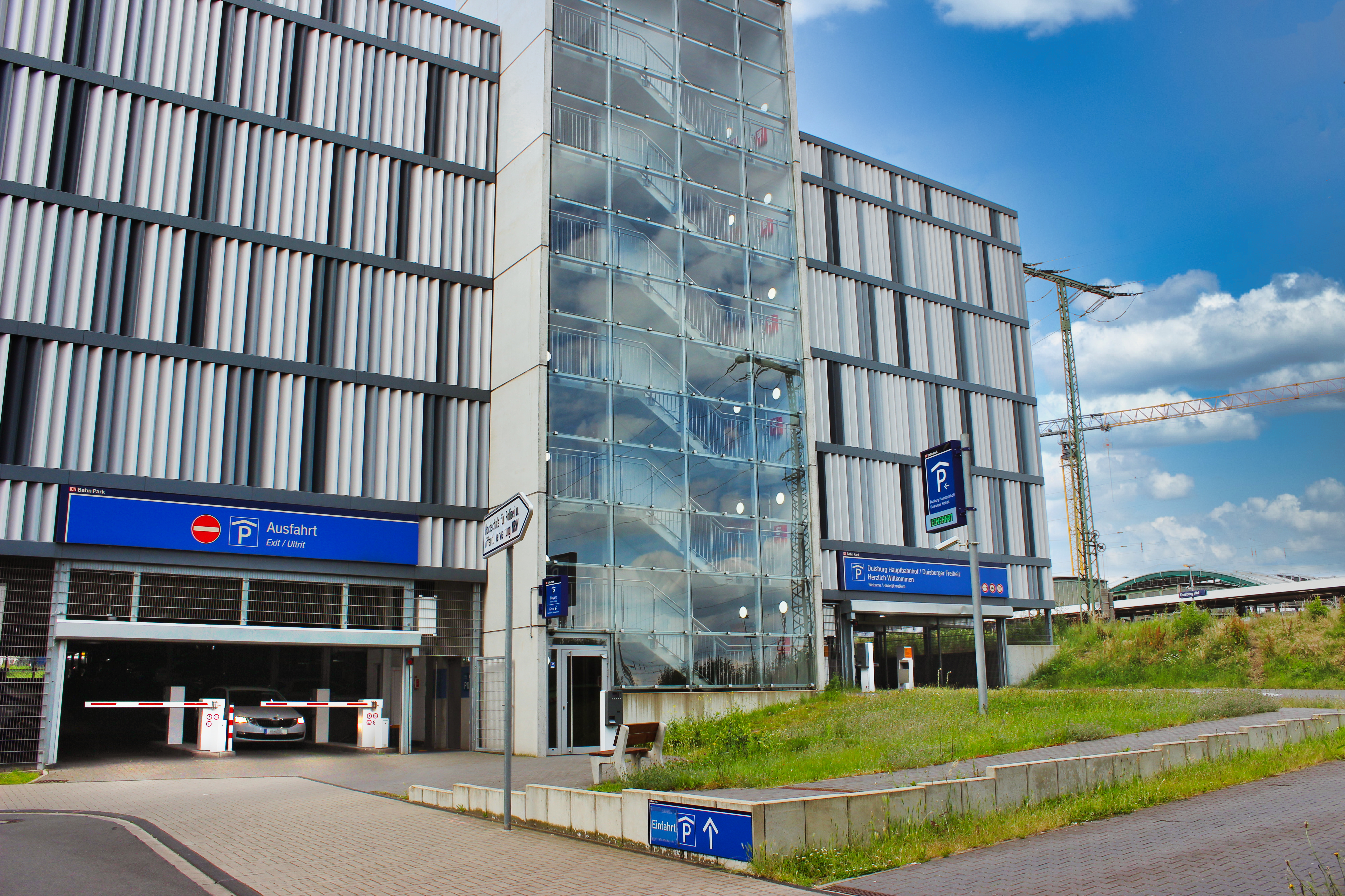 Es ist ein Bild des Parkhauses am Duisburger Hauptbahnhof der Contipark International Parking GmbH und DB Bahnpark dargestellt. Auf der linken Seite sieht man die Ausfahrt, auf der rechten Seite die Einfahrt. Im Hintergrund sieht man einen blauen Himmel mit vereinzelten Wolken.