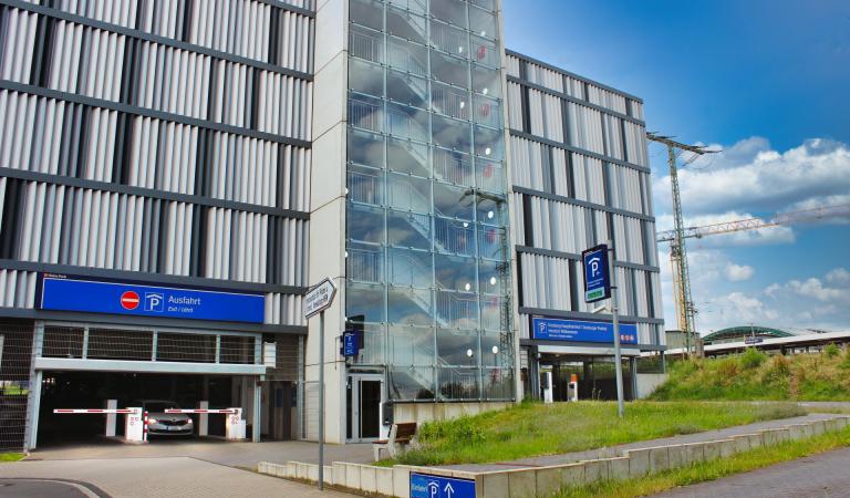 Obrázek parkoviště u hlavního nádraží v Duisburgu, které patří společnostem Contipark International Parking GmbH a DB Bahnpark. Na levé straně je vidět výjezd, na pravé straně vjezd. V pozadí vidíte modrou oblohu s rozptýlenými barvami
